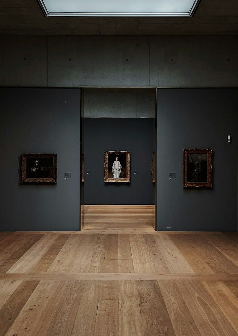 Link zur Galerie Ein neutrales Kunstmuseum mit dunklen Wänden und Holzboden, Blick auf Gemälde und Skulpturen.